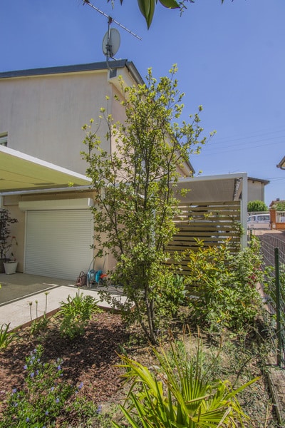 Maison moderne avec garage fermé blanc, jardin fleuri, jeune arbre et brise-vue en bois sous un ciel bleu d'été.