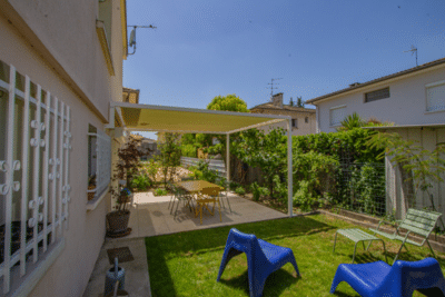 Jardin ensoleillé. Terrasse ombragée par une pergola, avec une table à manger. Pelouse verte et transats bleus modernes.