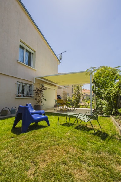 Maison beige avec volets et antenne. Jardin d'été lumineux, pergola jaune sur la terrasse. Mobilier extérieur et jeux bleus sur la pelouse.