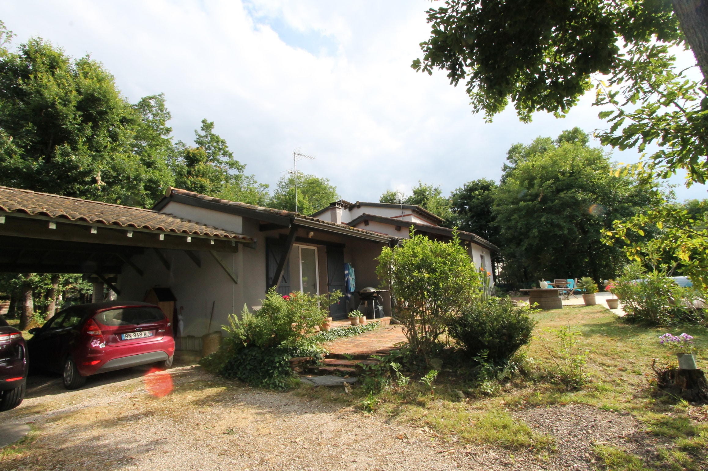 Maison de campagne provençale grise avec toit de tuiles, carport, voiture rouge. Terrasse et jardin fleuri, entourée de grands arbres.