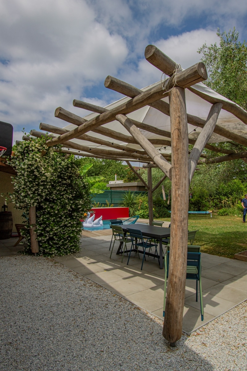 Terrasse avec pergola rustique en bois. Une table et des chaises colorées sont à l'ombre près du jasmin et d'une piscine extérieure.