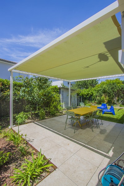 Pergola blanche et voile d'ombrage vert clair protégeant une terrasse carrelée. Mobilier de jardin jaune et vert dans un jardin ensoleillé.
