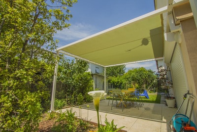 Pergola à toile tendue beige sur une terrasse ensoleillée, entourée d'un jardin luxuriant avec un calla lily blanc au premier plan.