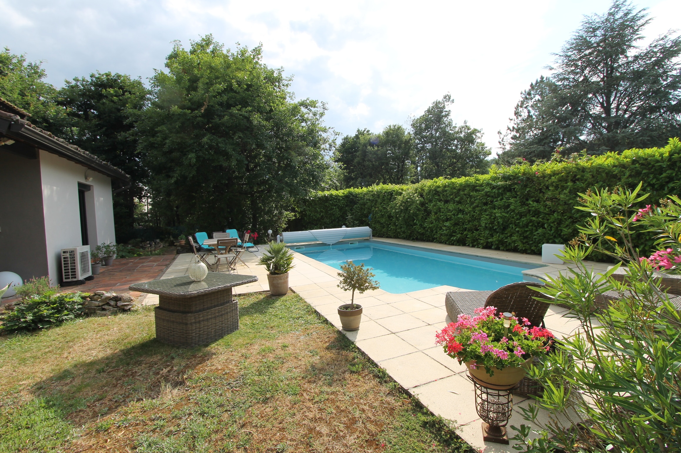 Longue piscine bleue couverte dans un jardin privé. Grande haie, arbres, terrasse claire et mobilier extérieur en rotin près de la maison.