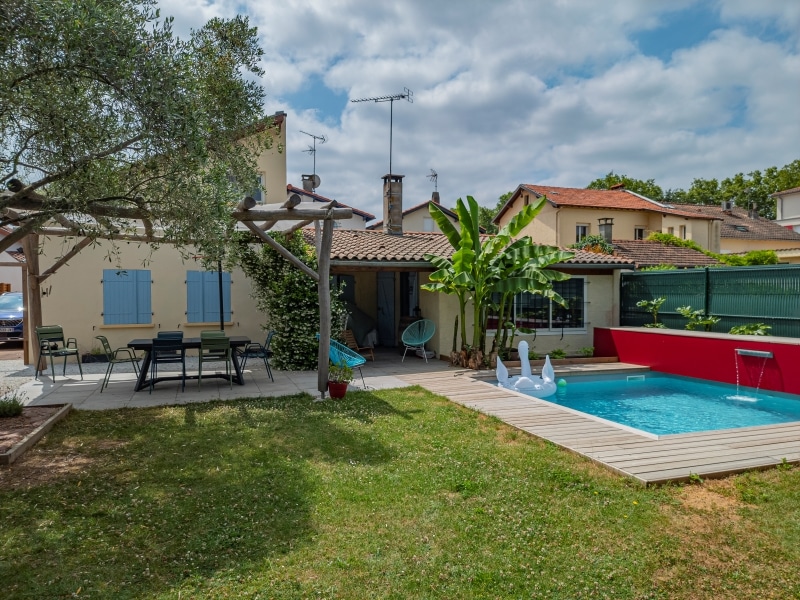 Piscine moderne avec mur rouge et cascade, terrasse en bois, mobilier de jardin ombragé par un olivier, et bananier devant une maison aux volets bleus.