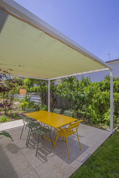 Pergola moderne blanche sur une terrasse carrelée. Grande table jaune et chaises de jardin vertes sous une toile d'ombrage, entourées de verdure.