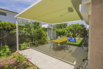 Terrasse carrelée sous pergola à toile jaune pâle. Grande table à manger extérieure, chaises vertes. Vue sur le jardin verdoyant et la pelouse.