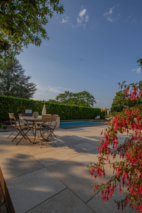 Aménagement de piscine en dalles de pierre claire, salon de jardin et fuchsia rouge sous un ciel bleu. Longue haie verte.