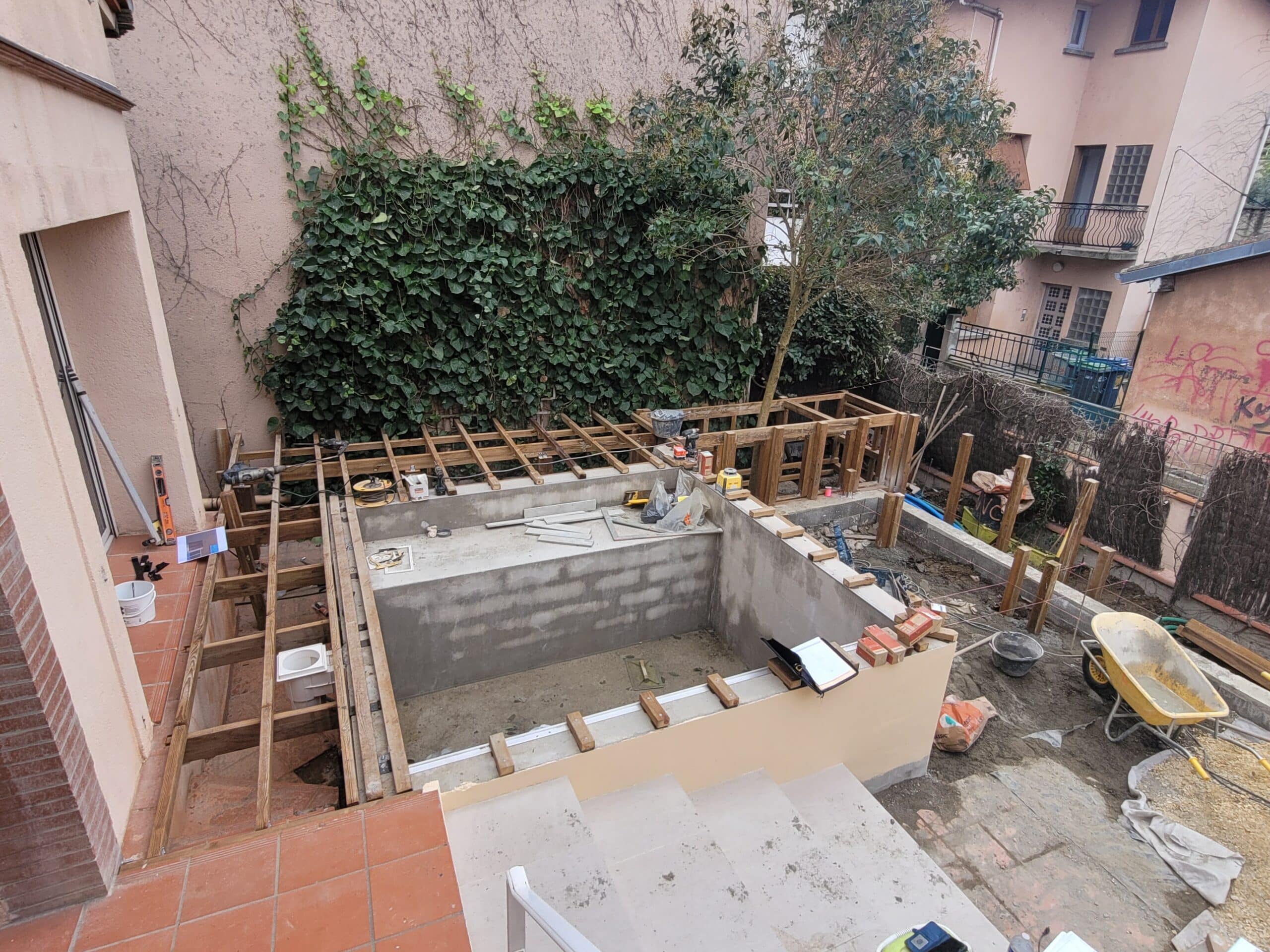 Construction d'une piscine en béton et terrasse bois dans un jardin urbain avec mur de lierre et matériel de chantier.