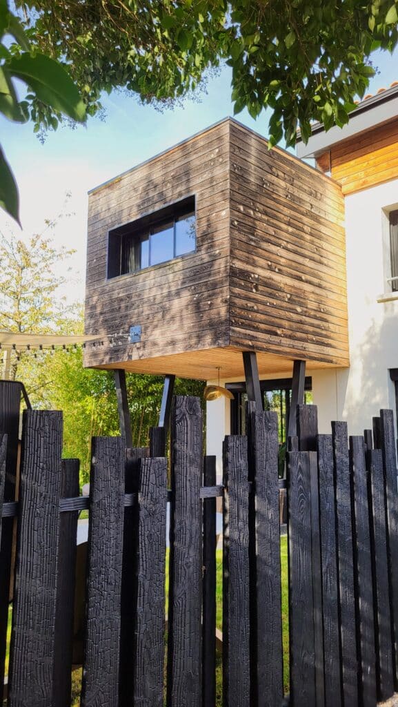 Extension moderne en bois sur pilotis et clôture en bois brûlé shou sugi ban dans un jardin ensoleillé.