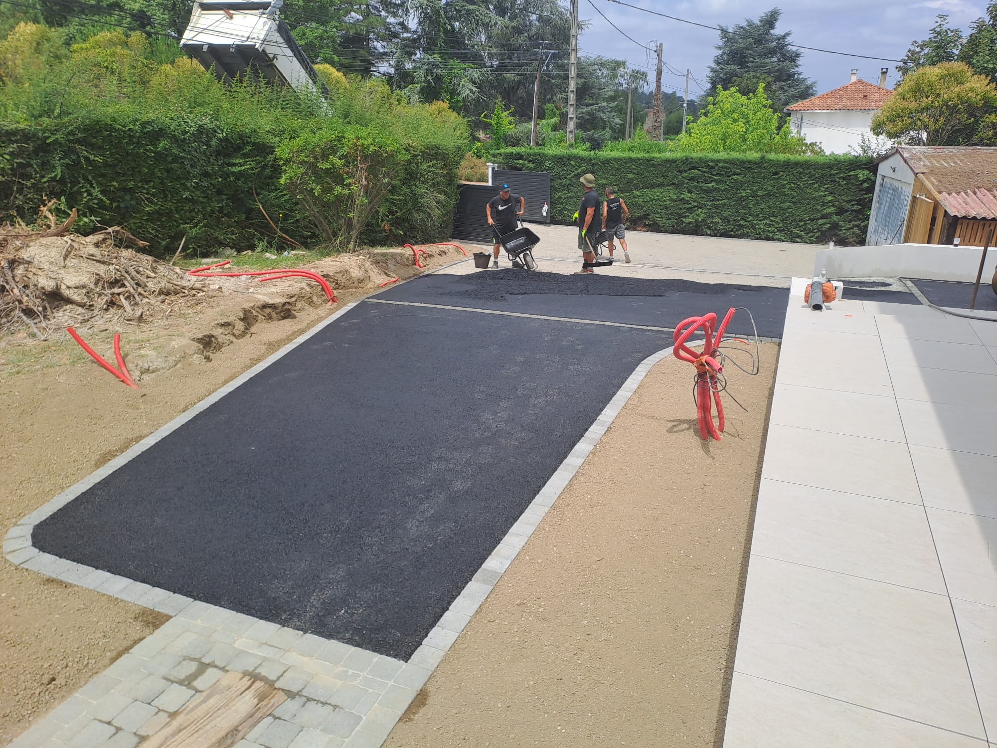 Ouvriers posant de l'enrobé noir sur une allée bordée de pavés lors d'un aménagement extérieur de maison.
