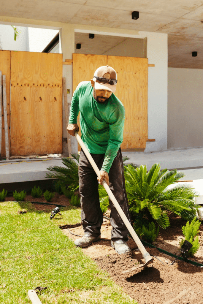 Paysagiste en chemise verte utilisant une houe pour préparer le sol près de plantes tropicales et d'une villa.