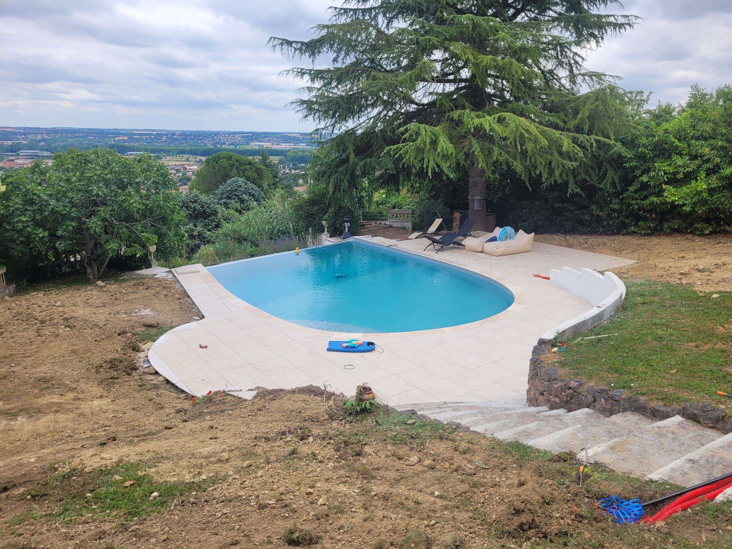 Piscine en forme de goutte, terrasse en dalles claires, grand cèdre et vue sur la vallée sous un ciel nuageux.