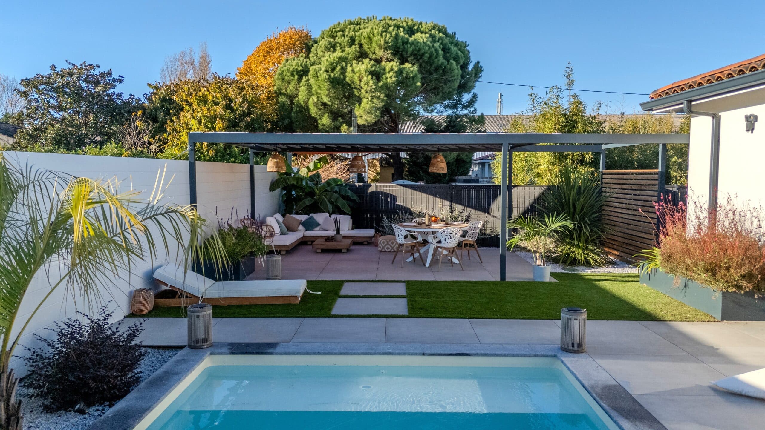 Aménagement extérieur moderne avec piscine, pergola grise, salon de jardin et coin repas sous un ciel bleu dégagé.