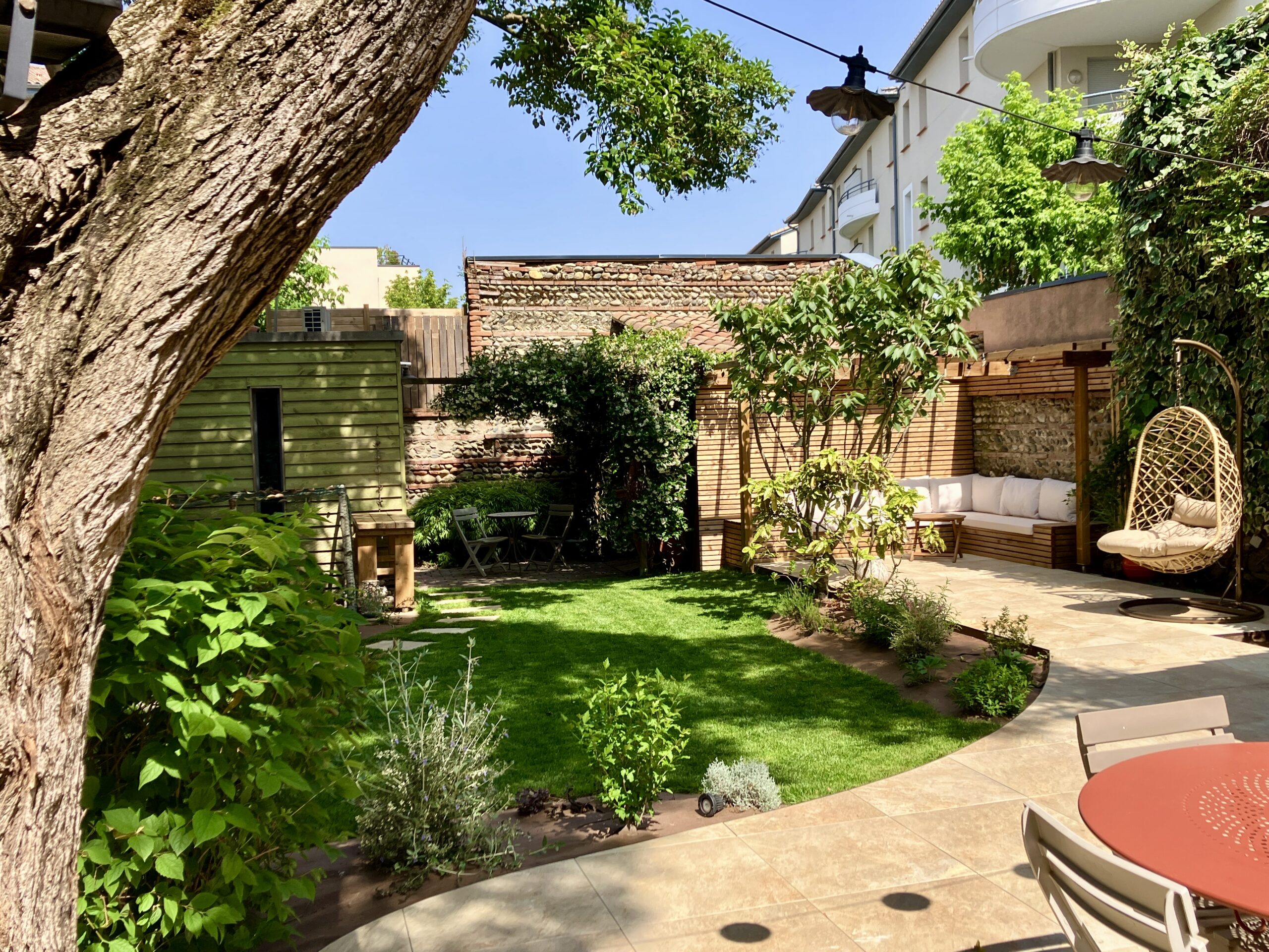 Jardin moderne avec pelouse, terrasse en pierre, banquette sous pergola bois et fauteuil suspendu relaxant.