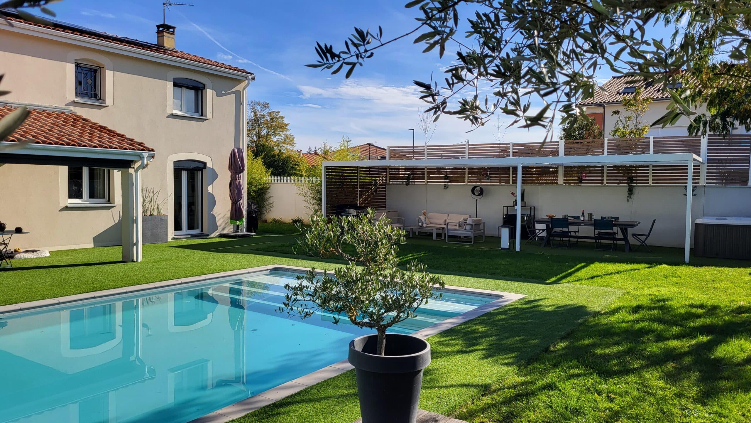 Jardin moderne avec piscine, pelouse synthétique, pergola aménagée et olivier en pot sous un ciel bleu.