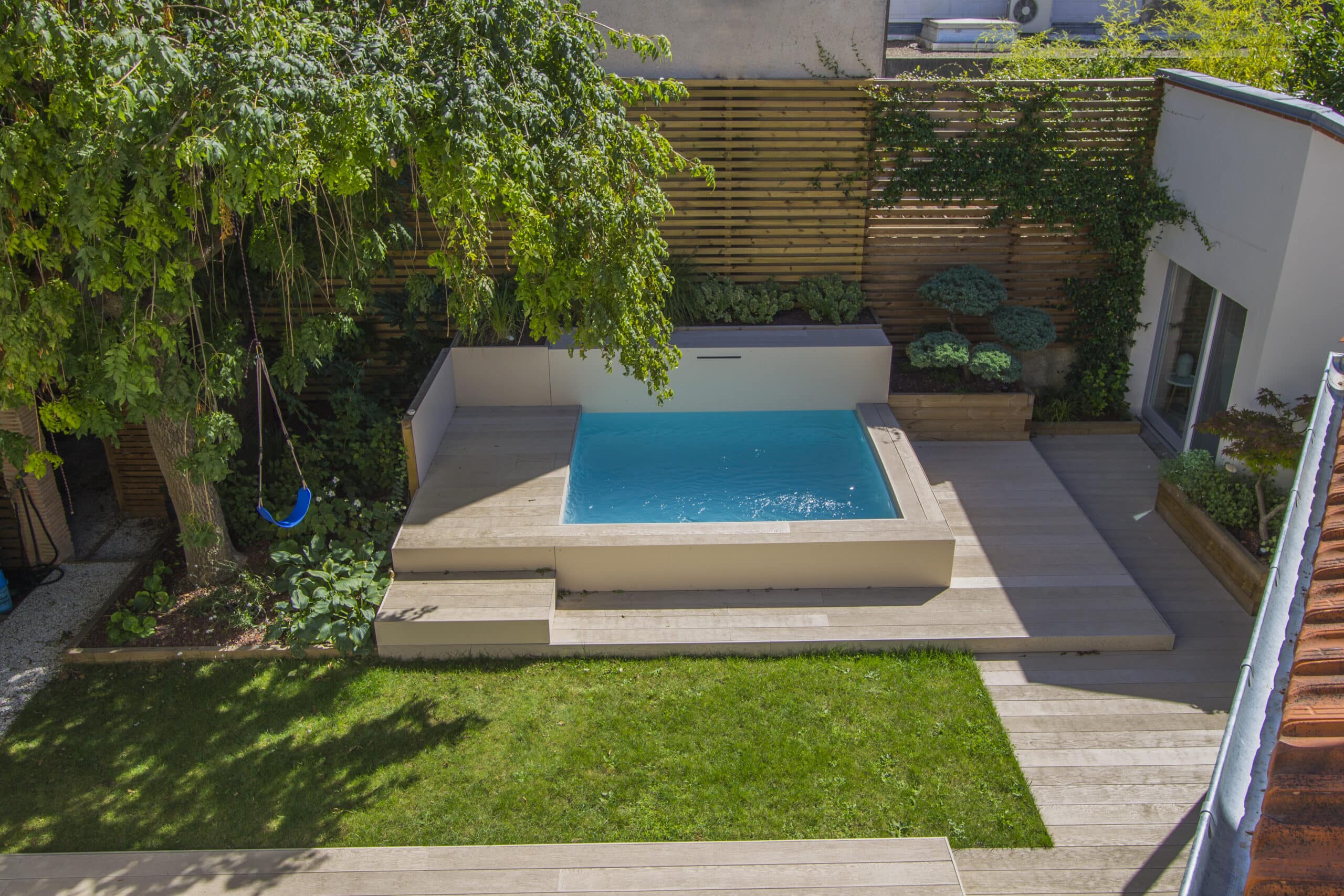 Petite piscine carrée sur terrasse en bois avec balançoire bleue sous un arbre et jardin paysager moderne.