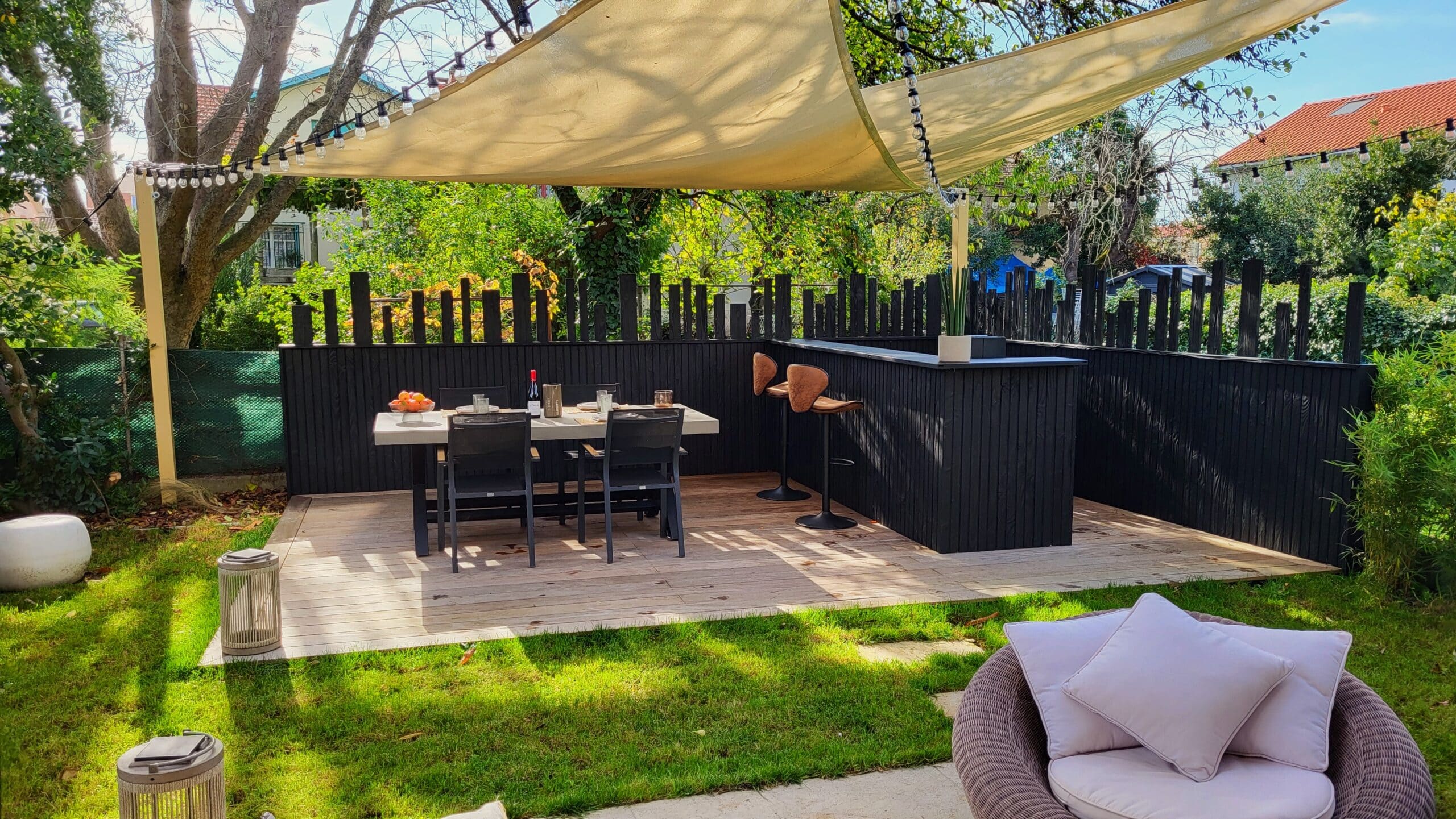 Aménagement d'une terrasse moderne avec voile d'ombrage, table à manger, bar en bois noir et guirlandes lumineuses.