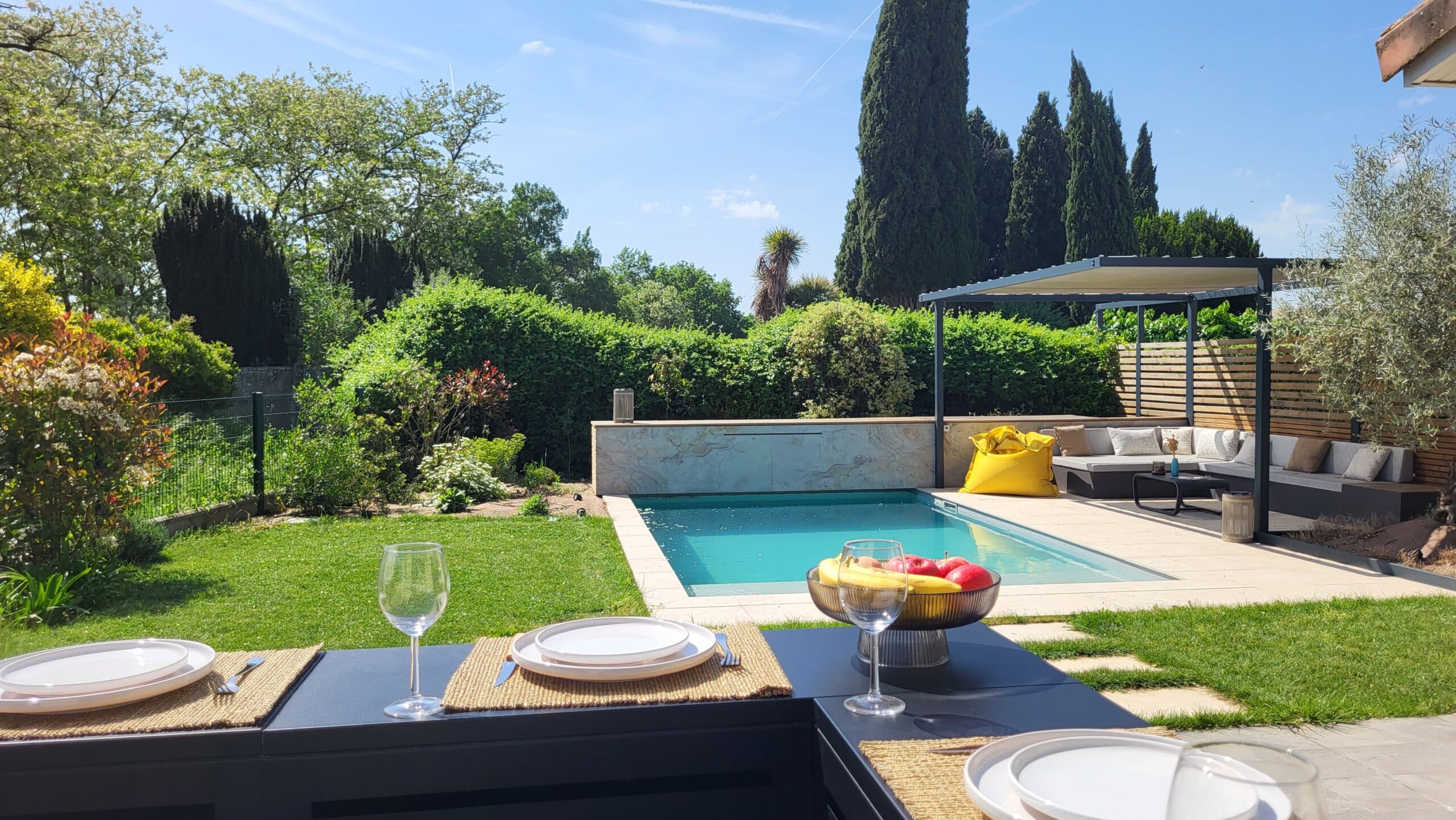 Table dressée devant une piscine moderne et un jardin paysager ensoleillé avec salon de terrasse.