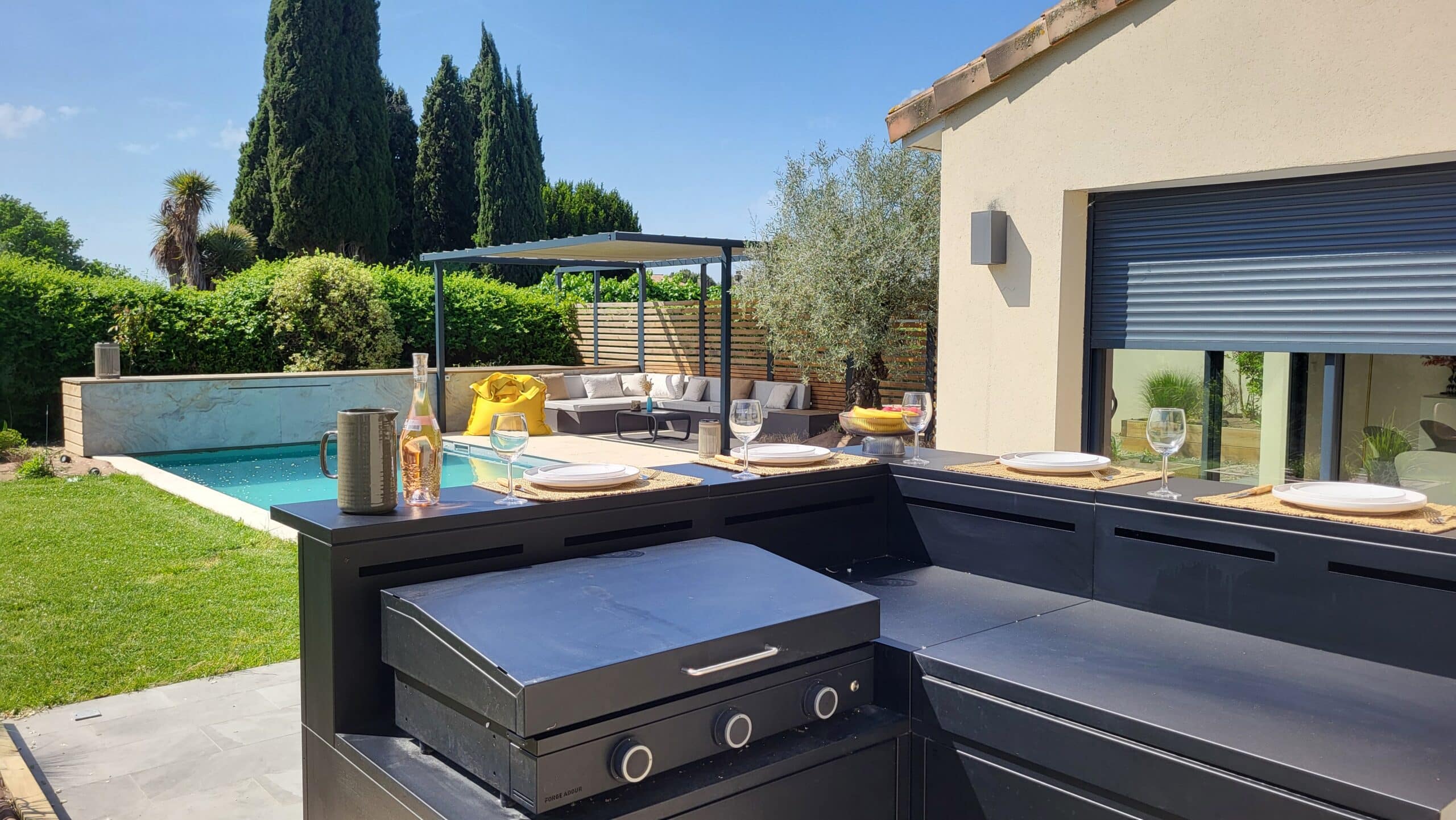 Cuisine d'été moderne avec plancha Forge Adour, table dressée, vue sur piscine et salon de jardin.