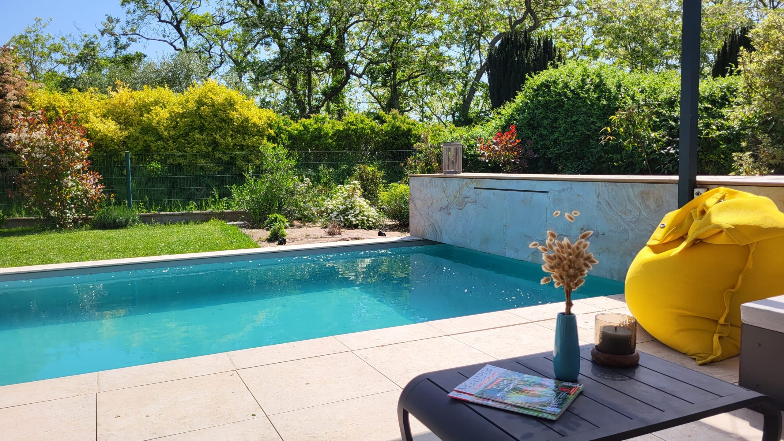Piscine bleue, terrasse avec pouf jaune et table basse devant un jardin verdoyant et ensoleillé.