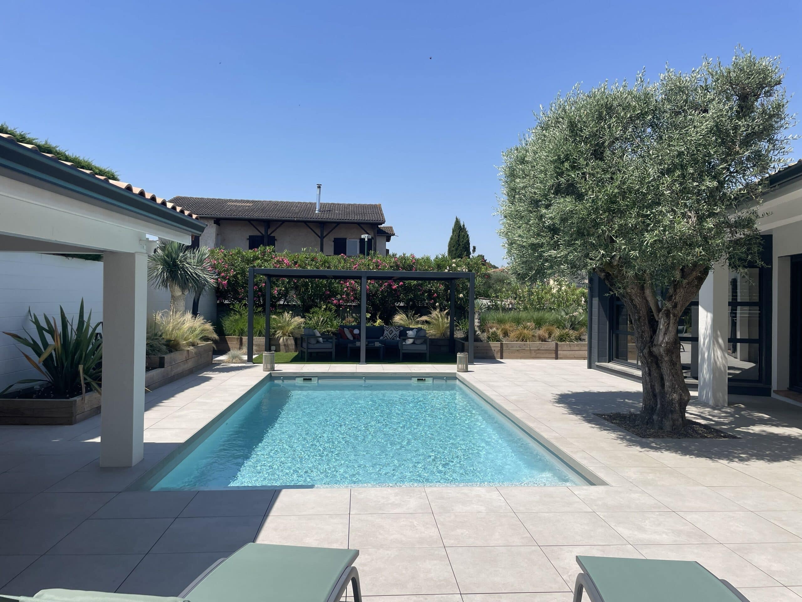 Piscine moderne, terrasse grise, olivier et pergola avec salon de jardin sous un ciel bleu dégagé.