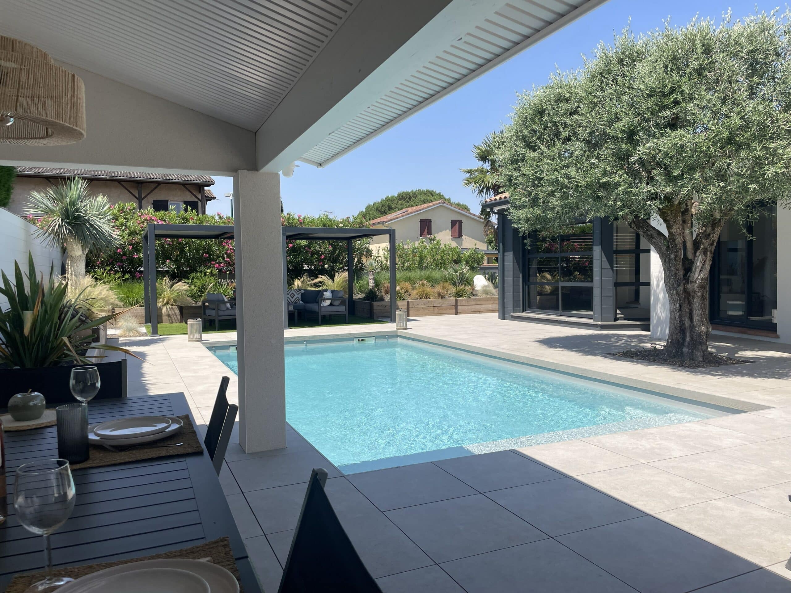 Piscine rectangulaire, terrasse carrelée, pergola et grand olivier sous un ciel bleu ensoleillé.