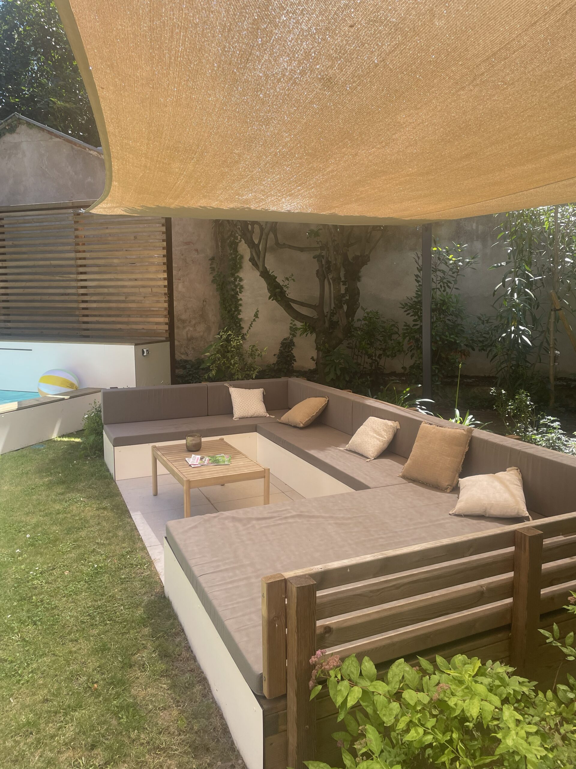 Salon de jardin gris sous voile d'ombrage beige avec table basse en bois et piscine en arrière-plan.