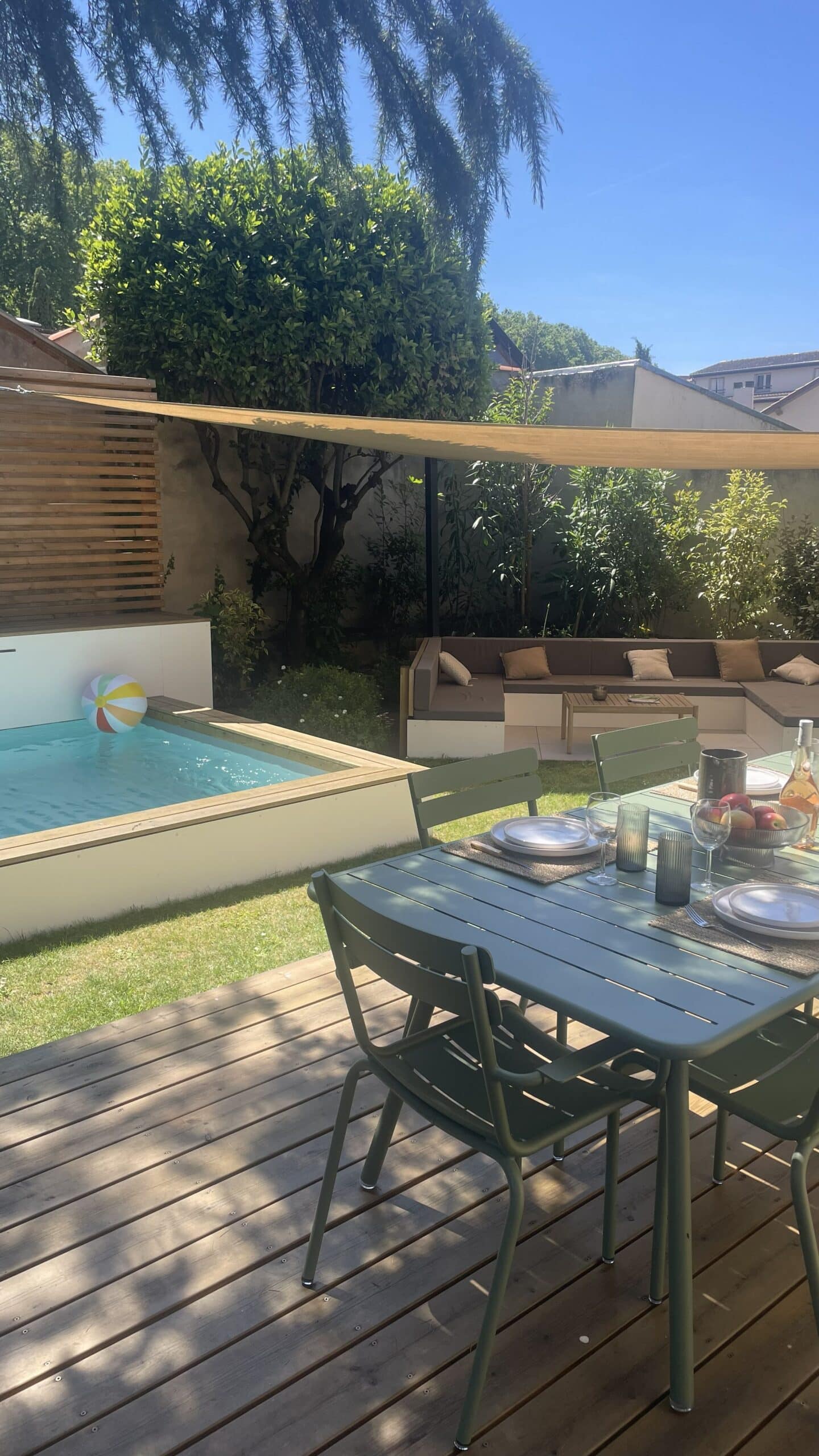 Terrasse en bois avec table dressée, petite piscine et salon de jardin sous voile d'ombrage ensoleillé.