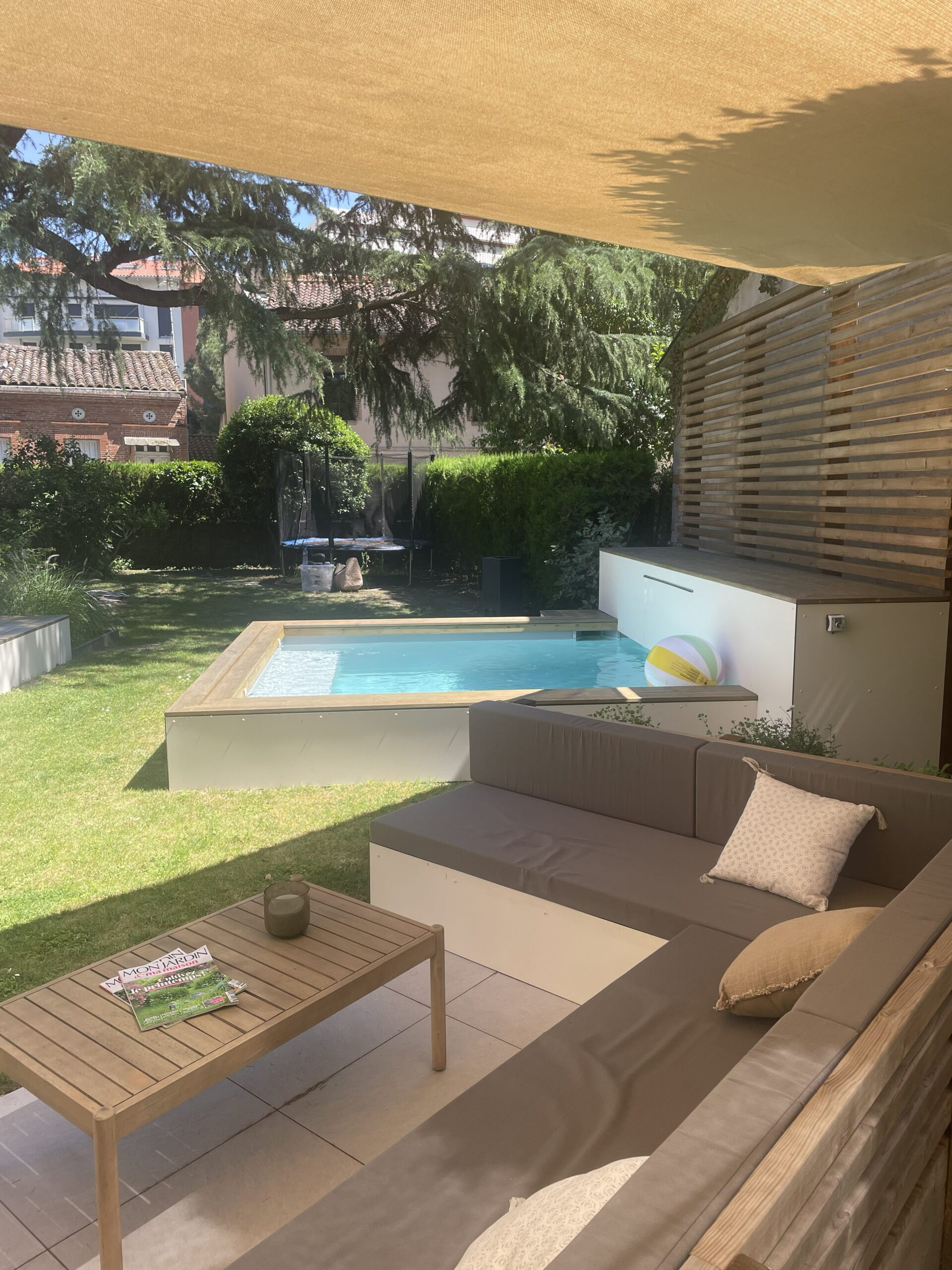 Terrasse moderne avec canapé d'angle, petite piscine de jardin et mobilier en bois sous un voile d'ombrage.