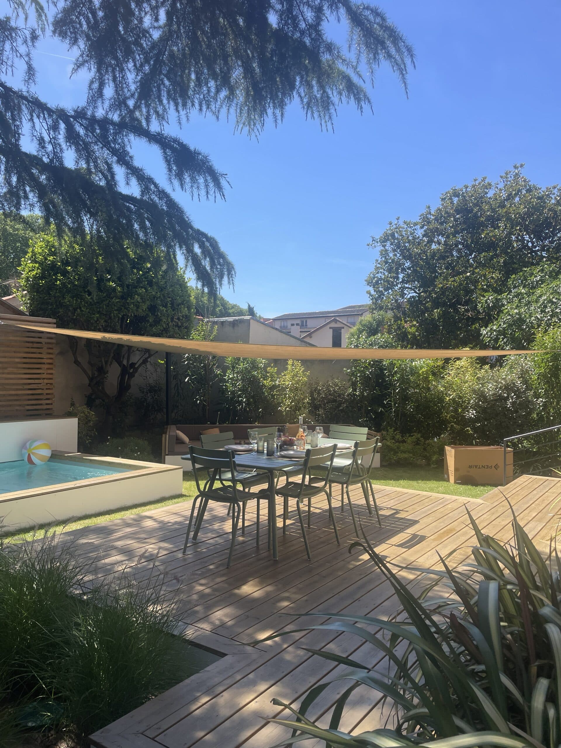 Terrasse en bois avec table à manger, piscine, voile d'ombrage et jardin verdoyant sous un ciel bleu.
